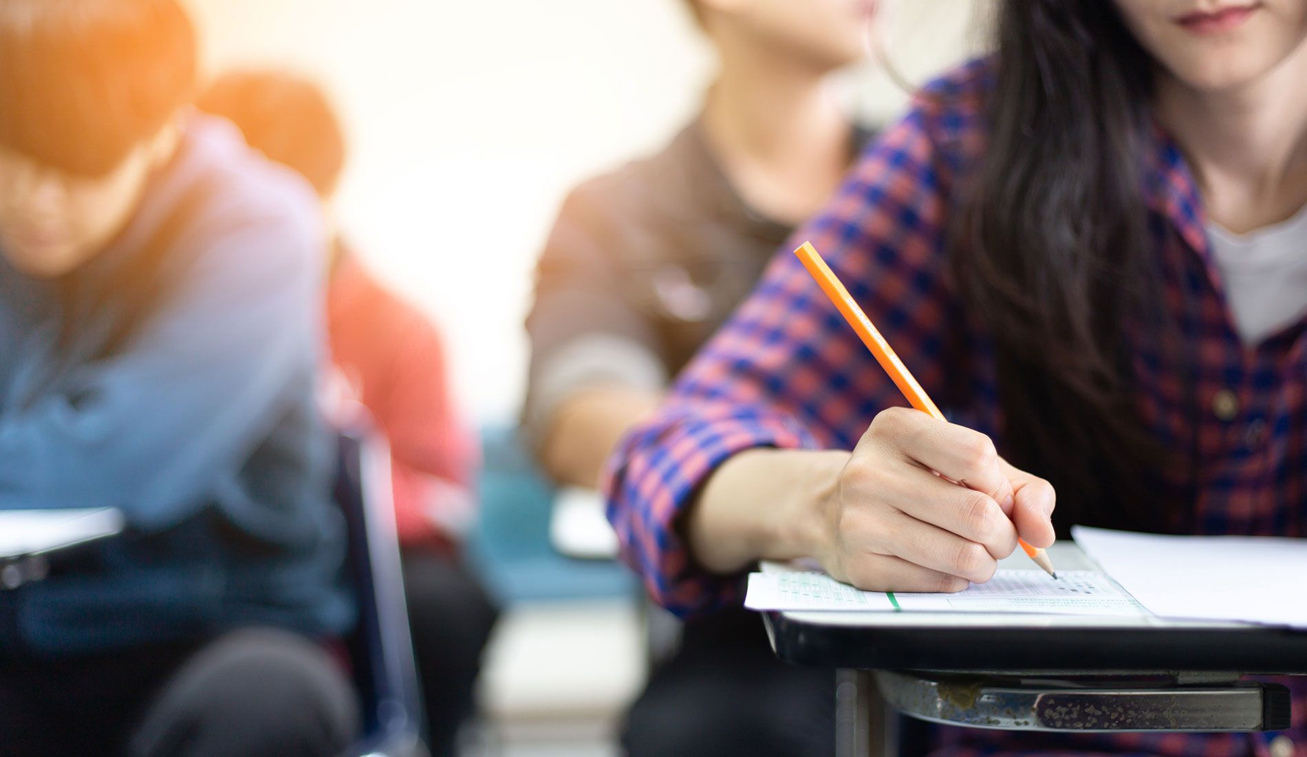 Fotka na detail studenta sedícího v lavici, jak píše něco tužkou na papír